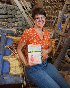 Mélissa Fialon présentant son livre "En attendant l'hiver" assise sur un char de paille. Derrière on voit un mur de pierres, le char est de couleur bleu.