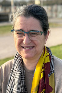 Sarah Oulion, souriante, portant une écharpe jaune, bordeaux avec des tons gris, noir, blanc. Le fond de cette image représente un chemin bordé par une pelouse de chaque côté. L'arrière-plan de l'image est flou.